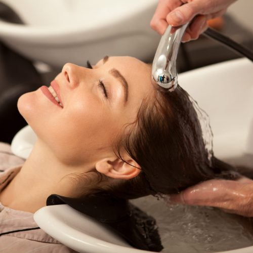 Happy beautiful woman smiling with her eyes closed while professional hairdresser washing her long healthy hair. Attractive woman enjoying relaxing hair treatment at beauty salon